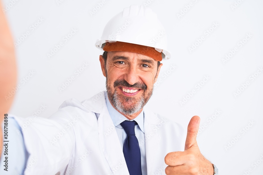 Middle age engineer man wearing helmet make selfie over isolated white background happy with big smile doing ok sign, thumb up with fingers, excellent sign Middle age engineer man wearing helmet make selfie over isolated white background happy with big smile doing ok sign, thumb up with fingers, excellent sign