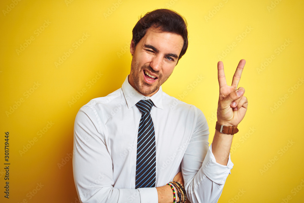 Young handsome businessman wearing elegant shirt and tie over isolated yellow background smiling with happy face winking at the camera doing victory sign. Number two. Young handsome businessman wearing elegant shirt and tie over isolated yellow background smiling with happy face winking at the camera doing victory sign. Number two.