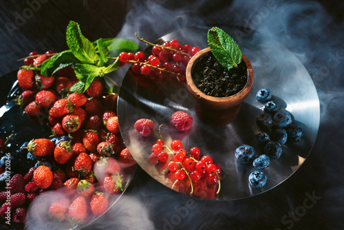 Tastes of hookah tobacco concept: raspberry, strawberry, blackberries, cranberries, black currants, wild berries. Hookah and fruits on dark wood table with shisha smoke clouds photo