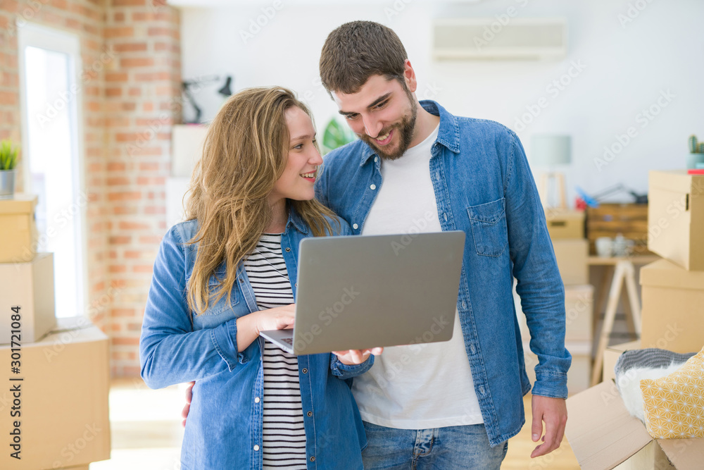 Young couple using computer laptop standing on a room around cardboard boxes, happy for moving to a new apartment Young couple using computer laptop standing on a room around cardboard boxes, happy for moving to a new apartment