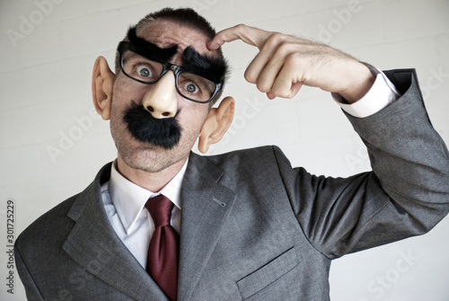 Confused businessman wearing a Groucho Marx disguise of glasses with thick eyebrows and mustache scratching his head looking at the camera