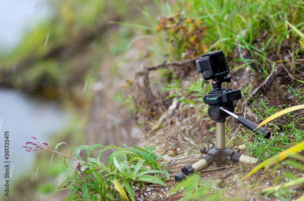 Action camera on a small tripod stands in the grass of the river under the flying snow flakes. Action camera on a small tripod stands in the grass of the river under the flying snow flakes.