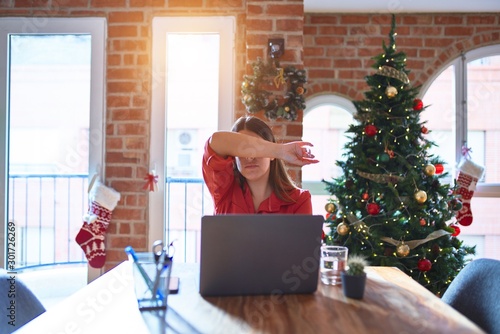 Beautiful woman sitting at the table working with laptop at home around christmas tree covering eyes with arm, looking serious and sad. Sightless, hiding and rejection concept