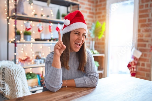 Young beautiful woman wearing christmas hat sitting at the table at home pointing finger up with successful idea. Exited and happy. Number one.