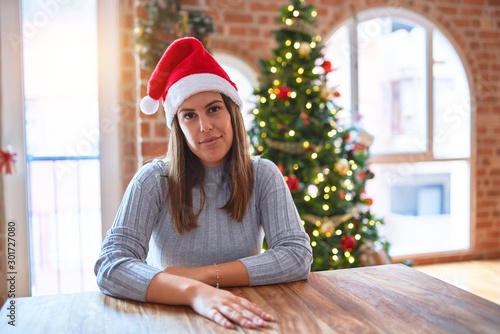Young beautiful woman wearing santa claus hat at the table at home around christmas decoration with serious expression on face. Simple and natural looking at the camera.