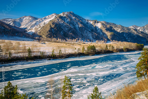 Majestic panoramic view of the valley covered with snow, turquoise river on the background of snow-capped mountains and clear blue sky. Sunny winter day in the mountains.
