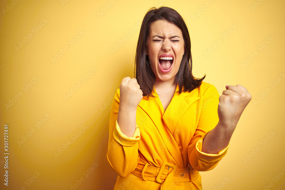 Young beautiful brunette woman wearing elegant dress over yellow isolated background very happy and excited doing winner gesture with arms raised, smiling and screaming for success. Celebration - obrazy, fototapety, plakaty 