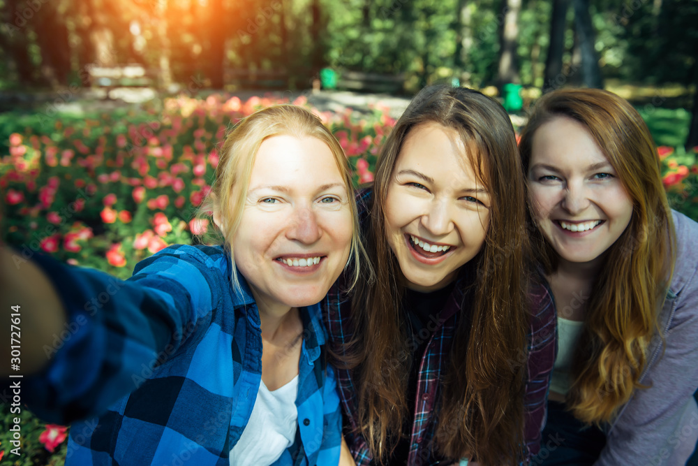 Happy young girls have fun laughing and taking selfies on a smartphone in the park on the background of green foliage and flowers. Blonde, brunette and girl with red hair, female friendship concept. Happy young girls have fun laughing and taking selfies on a smartphone in the park on the background of green foliage and flowers. Blonde, brunette and girl with red hair, female friendship concept.
