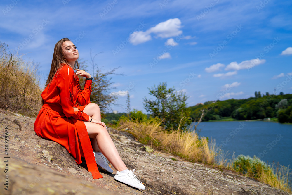 portrait of a beautiful young woman in red long dress. Lake view. Nature  background. Long hair. Nice makeup. Professional summer photoshoot.  Closeup. Girl sitting on a rock, long beautiful legs. Stock Photo |