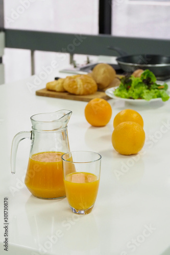 Glass and pitcher of orange juice on white table in kitchen with oranges, bun and salad for breakfast , focus at orange juice.