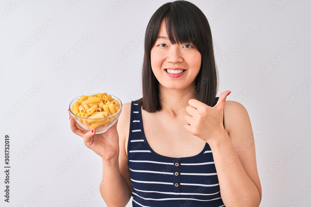 Young beautiful Chinese woman holding bowl with macaroni over isolated white background happy with big smile doing ok sign, thumb up with fingers, excellent sign Young beautiful Chinese woman holding bowl with macaroni over isolated white background happy with big smile doing ok sign, thumb up with fingers, excellent sign