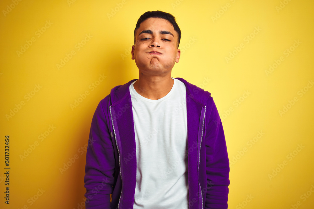 Young brazilian man wearing purple sweatshirt standing over isolated yellow background puffing cheeks with funny face. Mouth inflated with air, crazy expression. - obrazy, fototapety, plakaty 