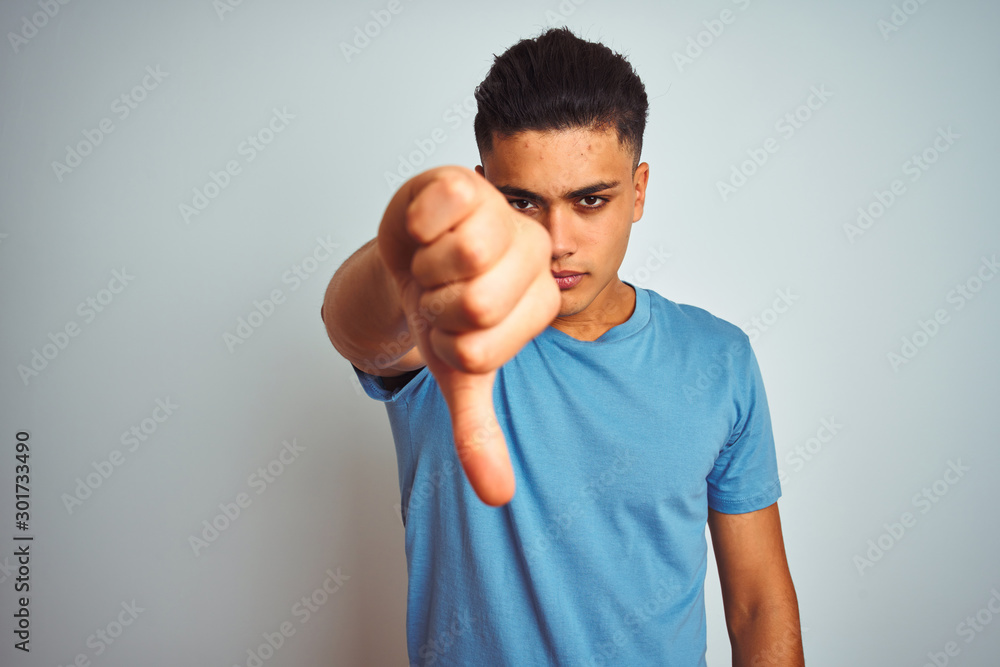 Young brazilian man wearing blue t-shirt standing over isolated white background looking unhappy and angry showing rejection and negative with thumbs down gesture. Bad expression. Young brazilian man wearing blue t-shirt standing over isolated white background looking unhappy and angry showing rejection and negative with thumbs down gesture. Bad expression.