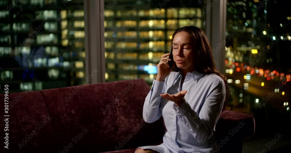 cute brunette sitting on the couch and fun talking on the phone at the window. she gestures and laughs. outside the window is dark and the lights of buildings glow. blurred lights of cars move cute brunette sitting on the couch and fun talking on the phone at the window. she gestures and laughs. outside the window is dark and the lights of buildings glow. blurred lights of cars move
