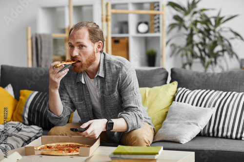 Obraz na plátně Portrait of middle-aged bearded man watching TV at home and eating pizza during