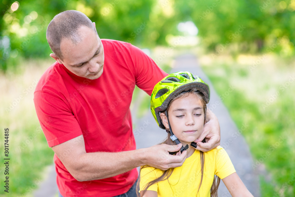 Naklejka premium Father puts a helmet on his daughter in summer park