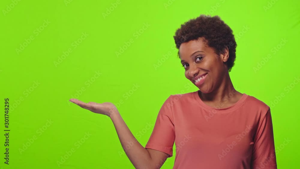 Studio shot of pleasant looking cheerful African woman raises palm near shoulder, holds copy space, smiles cheerfully, isolated over chroma key background, dressed in a casual t-shirt. Advertisement Studio shot of pleasant looking cheerful African woman raises palm near shoulder, holds copy space, smiles cheerfully, isolated over chroma key background, dressed in a casual t-shirt. Advertisement