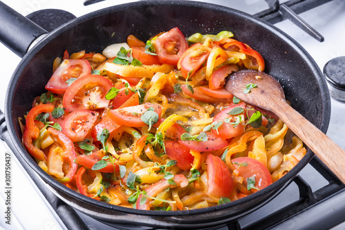 Sliced bell peppers with tomatoes and basil leaves are fried in a pan on a gas stove and mixed with a wooden spoon - cooking homemade sauce