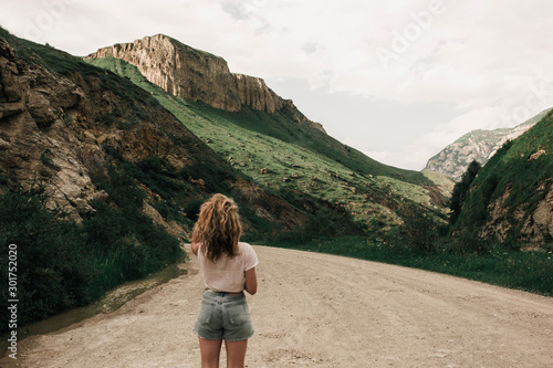 fashionable girl in white clothes standing on the road in the highlands . green grass and mountains