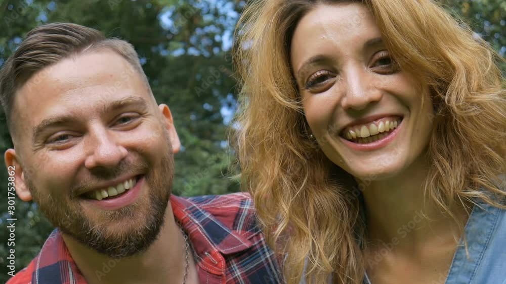 Lovely couple of bearded hipster boyfriend in red shirt and blond girlfriend wearing blue casual jeans blouse looking at the camera on green trees background Lovely couple of bearded hipster boyfriend in red shirt and blond girlfriend wearing blue casual jeans blouse looking at the camera on green trees background