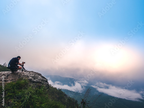 Nature photographer takes photos with camera on tripod at cliff. Dreamy foggy landscape. Morning sunrise blue misty in beautiful valley below.