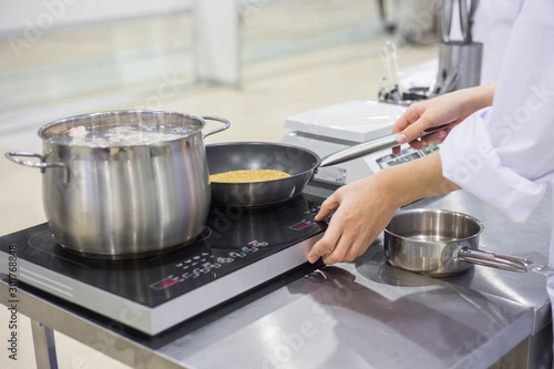 Chef cooking cereal, grain in frying pan with oil on electric stove at cuisine of restaurant. Professional cooking, catering, cookery, gastronomy and food concept