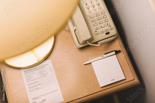 Close up photo of wooden bedside table. On the table golden lamp, telephone, notes with pen and questionnaire to improve the quality of service. Interior in hotel. Tourism concept. 