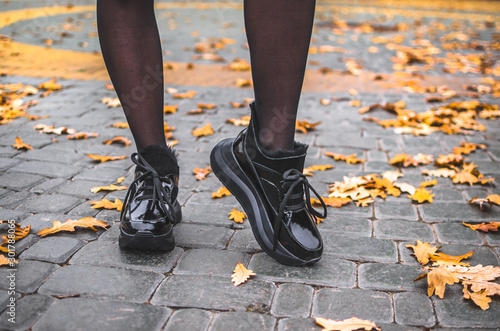 A girl walks in black shiny warm shoes on a tile with leaves in the city center in a park