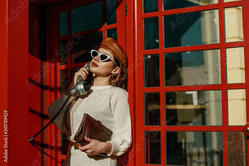 Outdoor autumn fashion portrait of young elegant woman wearing stylish  beret, sunglasses, white sweater, holding brown textured crocodile leather handbag, posing in red call box. Copy, empty space