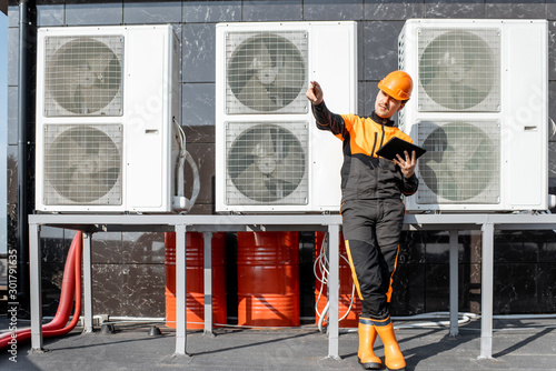 Egineer in protective workwear designing with digital tablet, while standing near the outdoor units of the air conditioner or heat pump photo