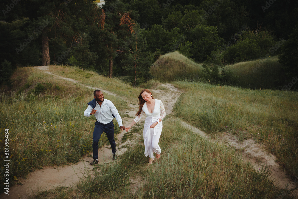 Fototapeta premium Cheerful mix raced couple enjoying vacation outside. Young man and woman walking up oldpark, holding hands and smiling