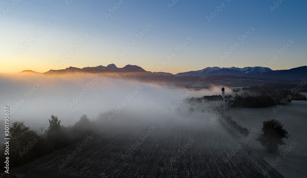 Beautiful foggy autumn morning in Bavaria - aerial view of Wilparting Church before Alps in Chiemgau