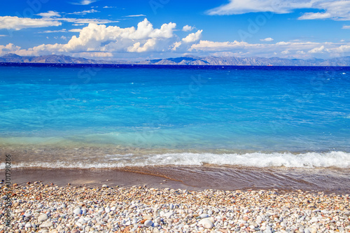 beach view with pebble beach sea, clouds and mountains in distance, Greek island.