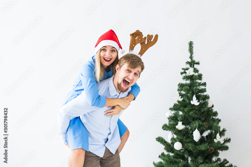 Obraz premium Funny man giving piggyback to his wife while they wearing Santa hats for Christmas holidays at home.