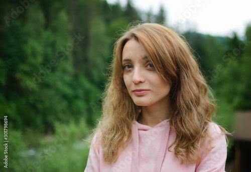 Attractive young calm blonde woman in pink clothes with long hair on the hill with green forest background during spring or early autumn in the mountains.
