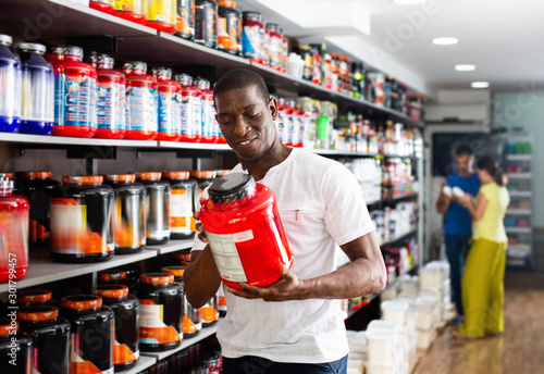 Focused muscular African man choosing sports nutrition products in shop, reading content label