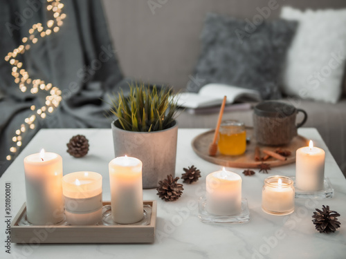 Cozy home  hygge  cosiness concept - burning white fragrance candles on white marble table near sofa with pillows and plaid. Winter decor  hot tea cup and honey