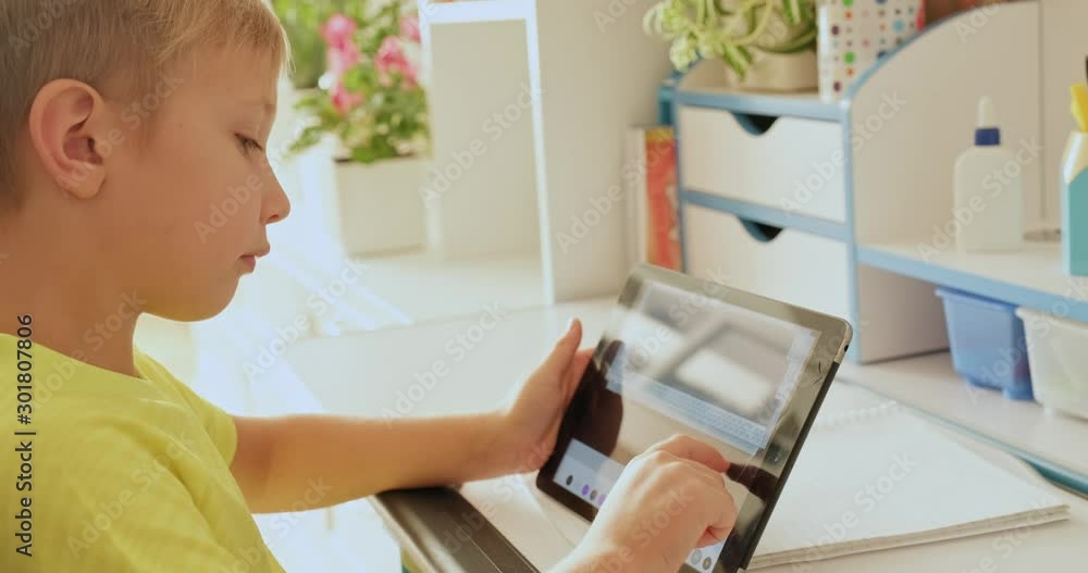 Schoolboy with tablet at home. Boy looks to the screen tablet. Caucasian kid holds tablet and typing text. Child spends time in the internet. Real time. Schoolboy with tablet at home. Boy looks to the screen tablet. Caucasian kid holds tablet and typing text. Child spends time in the internet. Real time.
