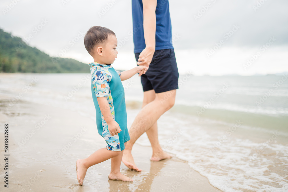 Asian toddler boy walking to the sea with his father.Dad holding hand his son for swim and play at the beach in Thailand. Asian toddler boy walking to the sea with his father.Dad holding hand his son for swim and play at the beach in Thailand.