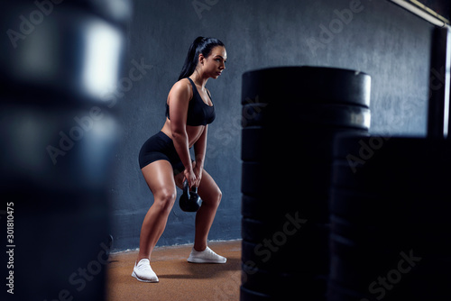 Woman fitness model make workout with weights - squat with kettlebell. Tanned sporty female  with black hair. Gym lifestyle.