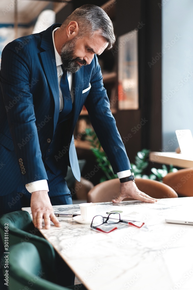 Stylish businessman standing near table in the office and reading documents Stylish businessman standing near table in the office and reading documents