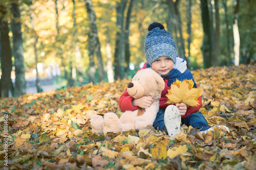 Little boy with his teddy bear friend sitting on the ground in the autumn park on sunset  nice back light.