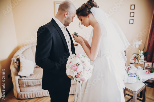 Photo Beautiful bride putting stylish boutonniere on groom suit, standing in room