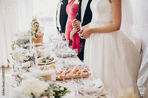 Stylish luxury centerpiece with floral decor, pink cloth, glasses and delicious food for wedding couple. Guests and bride with groom praying before eating. Catering at reception