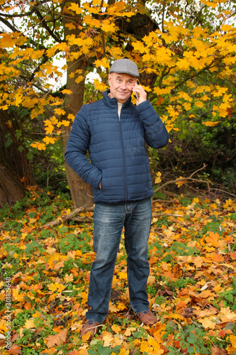 tinted photo of an elderly man in a gray cap and blue jacket, who is standing in an autumn park among maple trees, is phoning and smiling, a concept of telephone communication