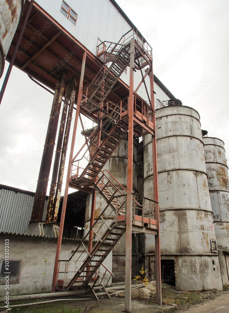 Technopathy, fragments of plant design and buildings. Metal staircases and columns of reinforced concrete products factory on the background of gray, cloudy sky. Technopathy, fragments of plant design and buildings. Metal staircases and columns of reinforced concrete products factory on the background of gray, cloudy sky.