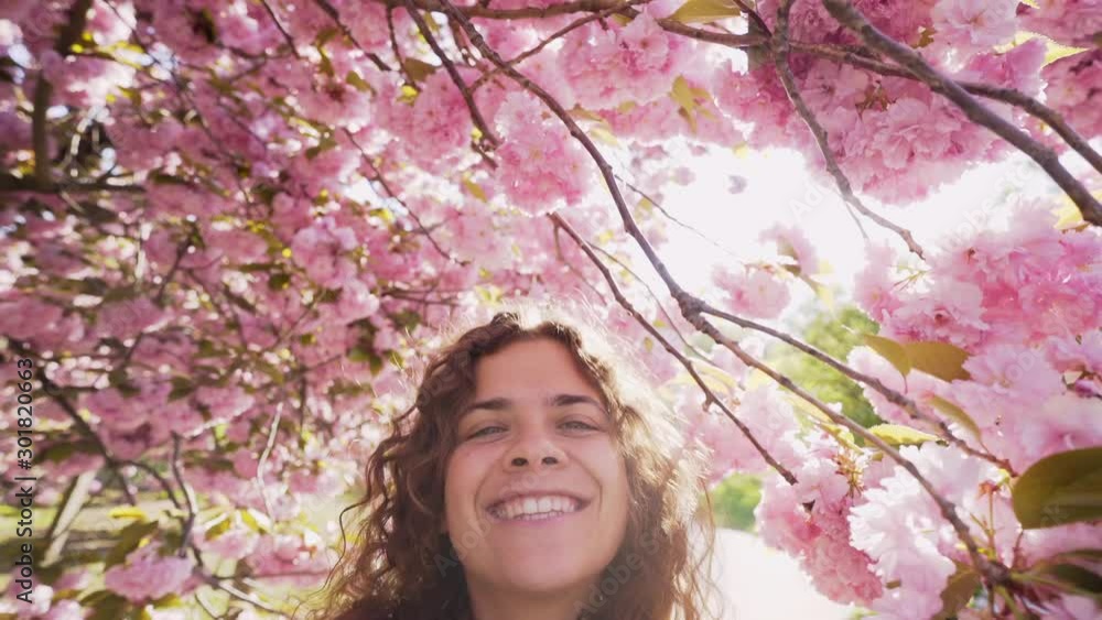 Young girl taking selfie under blossom cherry tree at backlit. Portrait playful woman, looking at camera, smiling and have fun while standing among branches covered flowers in bright sun light Young girl taking selfie under blossom cherry tree at backlit. Portrait playful woman, looking at camera, smiling and have fun while standing among branches covered flowers in bright sun light