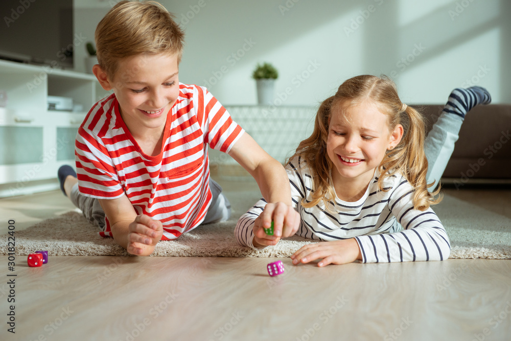 Portrait of two cheerful children laying on the floor and playing with colorful dices - obrazy, fototapety, plakaty 