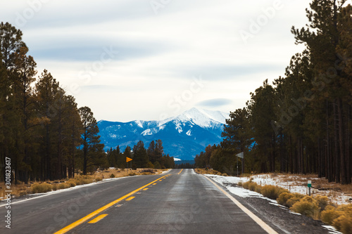 Beautiful America. Freeway leading towards snow covered Sanfrancisco Peak, in Flagstaff, Northern Arizona. Winter Travel. Lenticular cloud in the sky. Pine trees by the road sides, snow on road. 