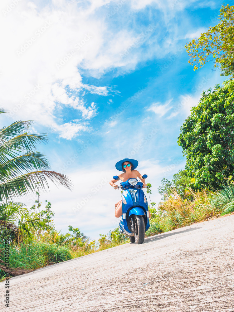 Obraz premium Crazy funny woman with flying hair riding a motorbike on a blue sky and green tropics background. Young bizarre girl with dark hair in sunglasses on a blue scooter in vintage style racing downhill